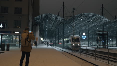 Hombre-Tomando-Video-Móvil-Del-Tren-Saliendo-De-La-Estación-Por-La-Noche-Helsinki