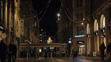 Calle-Con-Gente-Caminando-Y-Pasando-El-Tranvía-Por-La-Noche-Helsinki,-Finlandia