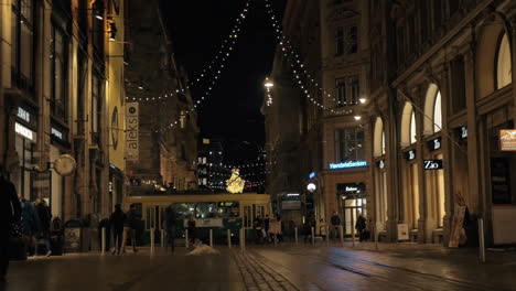 Gehende-Menschen-Und-Vorbeifahrende-Straßenbahn-In-Der-Nachtstraße-Mit-Weihnachtsbeleuchtung-Helsinki