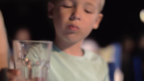 Niño-Aburrido-Bebiendo-Agua-En-Un-Café-Al-Aire-Libre-Por-La-Noche