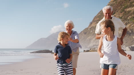 Grandparents-Chasing-Grandchildren-Along-Beach-On-Summer-Vacation