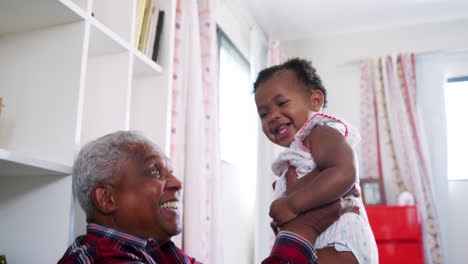 Grandfather-Lifting-Baby-Granddaughter-Into-The-Air-And-Smiling-At-Home