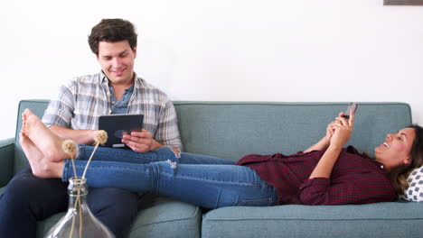 Couple-Relaxing-On-Sofa-At-Home-Using-Mobile-Phone-And-Digital-Tablet