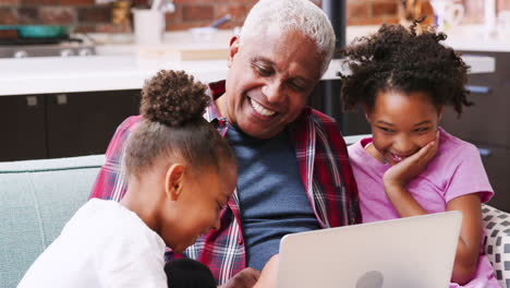 Grandfather-Sitting-On-Sofa-At-Home-With-Granddaughters-Using-Laptop