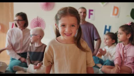 Ein-Glückliches-Vorschulmädchen-Mit-Blonden-Haaren-In-Einem-Gelben-T-Shirt-Schaut-In-Die-Kamera-Und-Lächelt-Vor-Dem-Hintergrund-Ihrer-Ersten-Unterrichtsstunde-Mit-Lehrern-In-Einführungsklassen.-Porträt-Eines-Kleinen-Mädchens,-Das-Vor-Dem-Hintergrund-Ihrer-Ersten-Unterrichtsstunde-Mit-Lehrern-In-Die-Kamera-Schaut