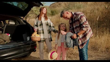 A-man-with-gray-hair-in-a-plaid-shirt-together-with-his-brunette-girlfriend-in-a-green-plaid-shirt-with-a-basket-of-food-and-their-little-daughter-are-preparing-to-go-on-a-picnic-in-a-steppe-area-with-dry-grass-in-summer