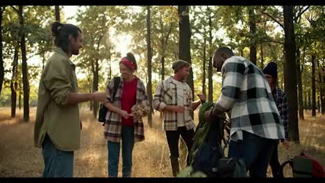 A-happy-group-of-people-on-a-hike-stop-at-a-halt-and-take-off-their-shoulders-their-backpacks-in-the-summer-forest.-A-brunette-man-in-a-green-jacket-communicates-with-his-friends-on-a-hike-and-tells-what-everyone-needs-to-do.-Distribution-of-responsibilities-during-the-trip