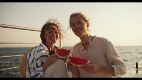 Portrait-of-a-happy-couple,-a-guy-in-light-clothes-and-a-girl-in-a-blue-and-white-shirt-are-holding-slices-of-watermelon-in-their-hands-and-looking-at-the-camera.-Guy-and-girl-on-a-picnic-on-the-pier-near-the-sea