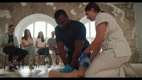 Close-up-shooting:-a-female-doctor-in-a-white-uniform-conducts-a-medical-first-aid-course-for-the-public,-who-listens-by-showing-artificial-respiration-on-the-mannequin-using-the-Ambu-resuscitation-bag-and-explains-his-actions,-a-Black-male-nurse-in-a-blue-uniform-performs-artificial-respiration-by-