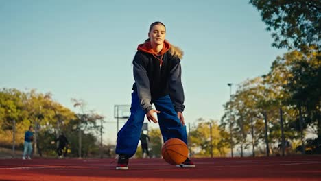 Below-is-a-portrait-of-a-blonde-girl-in-a-sports-uniform-and-blue-pants-who-is-hitting-an-orange-basketball-ball-off-the-red-floor-on-a-street-court-in-the-summer