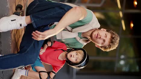 A-dad-with-curly-hair-in-a-Green-T-shirt-and-blue-jeans-sits-with-his-little-blond-son-in-a-black-safety-helmet-and-a-red-T-shirt-on-the-curb-in-a-park-near-a-building-and-looks-at-something-around-them