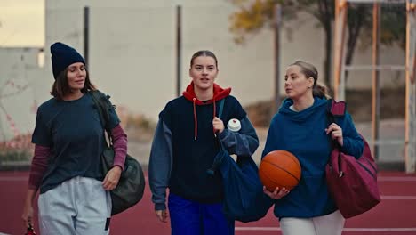 Tres-Chicas-Rubias-Seguras-De-Sí-Mismas,-Vestidas-Con-Ropa-Deportiva-Y-Con-Una-Pelota-De-Baloncesto-Naranja,-Caminan-Por-Una-Cancha-De-Baloncesto-Roja-En-Una-Mañana-De-Verano.-Chicas-Rubias-Felices-Van-A-Entrenar-Y-Jugar-Al-Baloncesto-En-Verano.