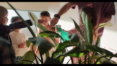 A-group-of-preschool-children,-together-with-their-teacher,-a-man-in-a-purple-T-shirt,-waters-an-indoor-flower-and-sprays-it-with-water-from-a-special-sprayer.-Biology-lesson-for-preschool-children