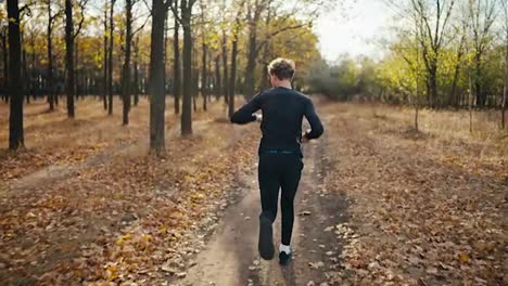 Rear-view-of-a-brunette-man-with-curly-hair-in-a-black-sports-uniform-runs-along-an-earthen-path-and-looks-back-at-his-watch-while-jogging-in-the-morning-in-an-autumn-forest-with-fallen-leaves