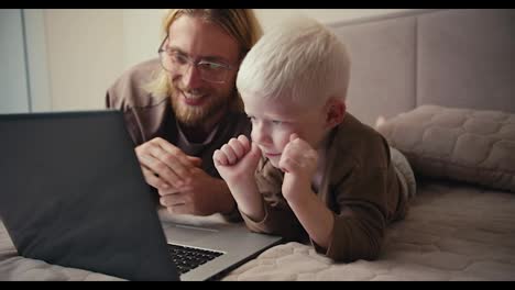 Happy-blond-man-in-glasses-with-a-beard,-together-with-his-little-albino-son-with-blue-eyes,-greets-and-waves-to-his-relatives-via-video-conference.-A-happy-albino-boy-with-white-hair-walks-into-a-video-conference-with-his-dad-and-waves-to-the-participants