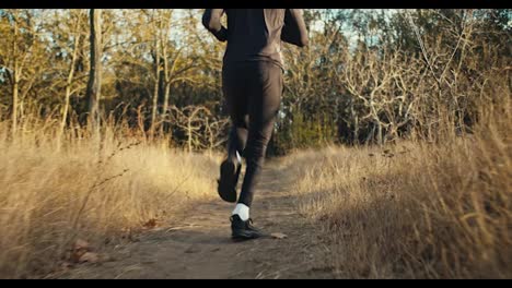 Rear-view-of-a-man-in-a-black-sports-uniform-in-black-sneakers-runs-along-a-narrow-path-among-dried-grass-in-the-autumn-forest-in-the-morning.-Morning-jog-in-the-park-in-autumn