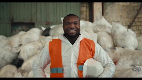 Close-up-portrait-of-a-happy-and-confident-man-with-Black-skin-in-a-white-protective-uniform-and-an-orange-vest-who-is-posing-near-a-pile-of-recycled-cellophane-and-plastic-at-a-waste-processing-and-sorting-plant