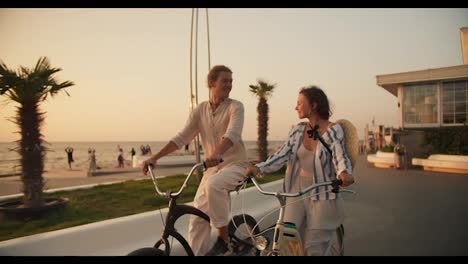 Front-view-of-a-guy-and-a-girl-riding-bicycles-and-looking-at-each-other-on-the-beach-on-which-palm-trees-grow-near-the-sea-in-summer-at-dawn.-Bike-ride-on-the-beach-in-the-morning