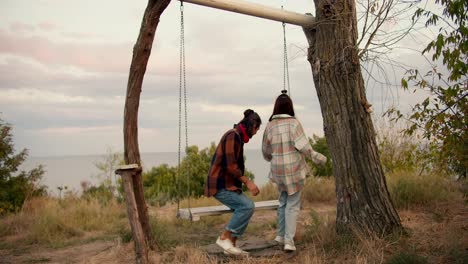 Shooting-from-behind:-A-guy-and-his-girlfriend-in-checkered-shirts-sit-on-a-wooden-swing-and-start-swinging.-Rest-in-the-country-by-the-sea