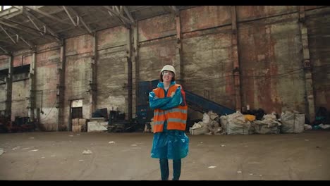 Close-up-portrait-of-a-brunette-girl-in-a-blue-protective-uniform-and-an-orange-vest-who-stands-in-the-huge-hall-of-a-waste-processing-plant-and-smiles