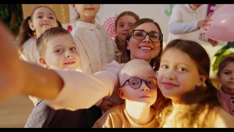 A-happy-teacher-with-a-bob-hairstyle-in-glasses-and-a-white-shirt-takes-a-selfie-with-her-students,-preschool-children-in-a-club-for-preparing-children-for-school