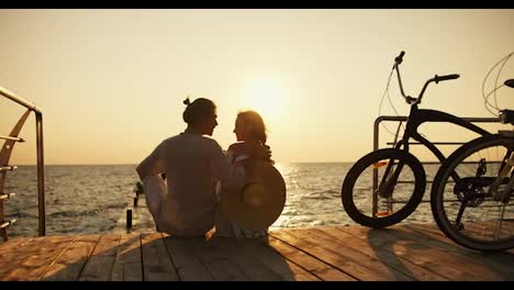 Zoom-out-guy-and-girl-sitting-on-the-pier-on-the-beach-opposite-the-sunrise-and-sea.-Happy-couple,-a-guy-in-light-clothes-and-a-girl-with-a-Straw-Hat,-arrived-on-bicycles-at-Sunrise-and-are-sitting-near-the-sea