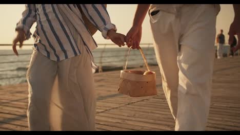 Close-up-shot-of-a-guy-in-white-pants-and-a-girl-in-a-white-skirt-walking-along-the-beach-holding-hands-and-carrying-a-basket-of-apples.-Happy-guy-girl-walking-along-the-pier-near-the-sea-on-a-date