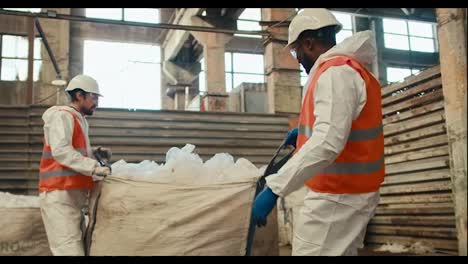 A-man-with-Black-skin-in-a-white-special-uniform-and-an-orange-vest-lays-out-and-sorts-cellophane-garbage-together-with-his-colleague-a-man-with-a-beard-in-a-white-helmet-at-a-waste-processing-and-sorting-plant