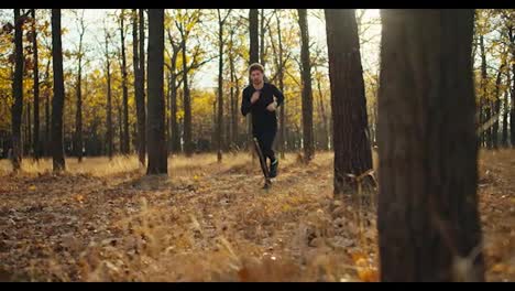 A-male-athlete-in-a-black-sports-uniform-runs-between-the-trees-covered-in-brown-leaves-and-dry-grass-in-the-fall-during-his-jog-on-a-sunny-day.-Happy-man-on-a-morning-jog-in-autumn