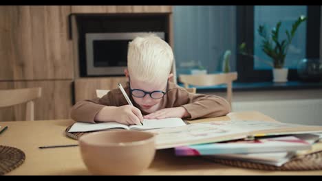 A-little-albino-boy-with-white-hair-in-blue-glasses-uses-a-pen-to-write-homework-in-his-notebook-in-the-kitchen-and-prepare-for-his-new-day-at-school-and-in-class.-A-little-albino-boy-writes-down-tasks-in-a-notebook-and-completes-them