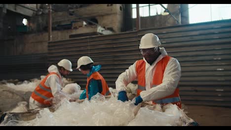 Ein-Mann-Mit-Schwarzer-Hautfarbe-In-Einer-Weißen-Schutzuniform-Und-Einer-Orangefarbenen-Weste-Sortiert-Und-Presst-Zusammen-Mit-Seinen-Kollegen-Plastikmüll-In-Zellophan-In-Einer-Abfallverarbeitungs--Und--sortieranlage.