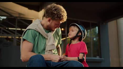 Happy-father-with-curly-hair-in-a-green-t-shirt-sits-near-his-little-son-dam-in-a-black-helmet-and-in-a-red-t-shirt-during-sunny-weather-in-the-evening-in-summer