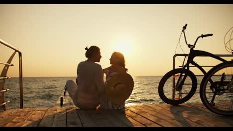 A-happy-couple,-a-guy-in-a-light-shirt-and-a-girl-in-a-straw-hat,-look-at-each-other-and-sit-on-a-beach-covered-with-boards-near-the-sea-at-sunrise.-A-guy-and-a-girl-came-to-the-beach-on-bicycles