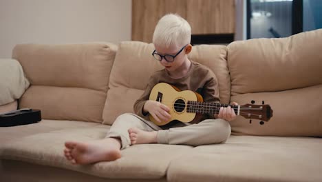 Kleiner-Albinojunge-Mit-Weißem-Haar-Und-Runder-Blauer-Brille-Spielt-Ukulele,-Während-Er-Abends-Auf-Einem-Cremefarbenen-Sofa-In-Einem-Modernen-Apartment-Sitzt.-Ein-Kleiner-Albinojunge-Mit-Weißem-Haar-Und-Blauer-Brille-Sitzt-Zu-Hause-Auf-Einem-Großen-Cremefarbenen-Sofa-Und-Spielt-Eine-Gelbe-Ukulele