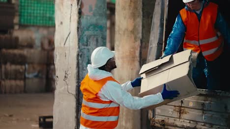 A-man-with-Black-skin-in-a-white-protective-uniform-in-an-orange-vest-accepts-waste-paper-given-to-him-by-a-brunette-girl-from-a-truck-and-stores-it-at-the-Large-Waste-Recycling-and-Sorting-Plant