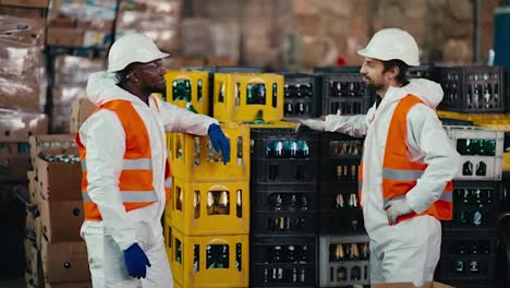 A-man-with-Black-skin-in-a-white-protective-uniform-in-an-orange-vest-communicates-with-his-male-colleague-with-a-beard-while-they-are-working-and-being-in-a-large-recycling-plant-for-plastic-and-glass-bottles