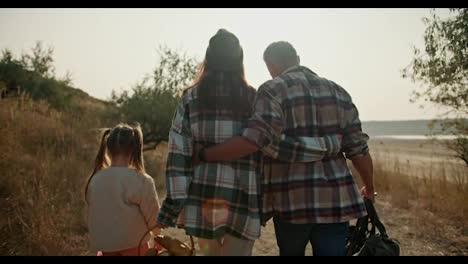 Rear-view-of-a-happy-couple,-a-brunette-girl-in-a-Green-checkered-shirt-and-a-Green-hat,-walks-and-hugs-with-her-husband-with-gray-hair-and-their-little-daughter-is-walking-next-to-them-on-a-picnic-along-the-road-with-dry-grass