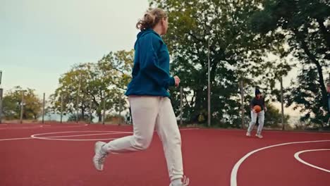 A-blonde-girl-in-a-turquoise-hoodie-and-sportswear-plays-basketball-on-the-Red-street-court-with-her-friends-in-the-morning-in-the-summer.-Blonde-girl-and-her-friends-play-basketball-in-the-morning-in-the-summer