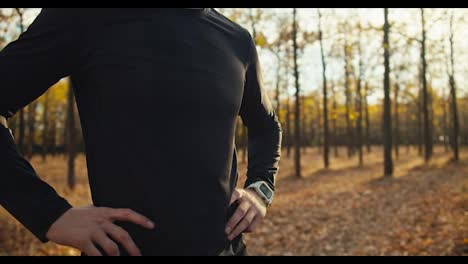 Portrait-of-a-confident-Happy-man-with-curly-hair-in-a-black-sports-uniform-and-a-beard-who-stands-in-the-middle-of-the-autumn-forest-and-looks-at-it-after-his-jog-on-a-sunny-autumn-morning