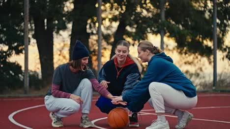 Three-happy-blonde-girls-in-sportswear-put-their-hands-together-and-raise-them-up-during-the-start-of-a-basketball-match-on-a-red-sports-court-in-the-morning