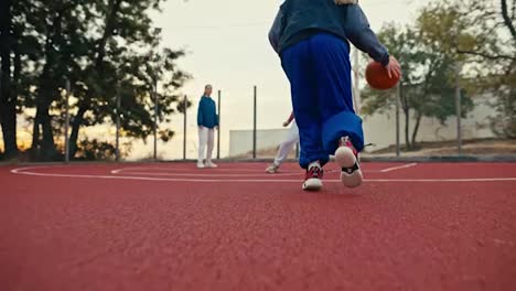 Filmada-Desde-Abajo,-Una-Chica-Rubia-Con-Pantalones-Azules-Y-Uniforme-Deportivo-Corre-Por-La-Cancha-Roja-Y-Acusa-A-Todos-Los-Oponentes-De-Lanzar-Una-Pelota-Naranja-A-Un-Aro-De-Baloncesto-En-Verano.