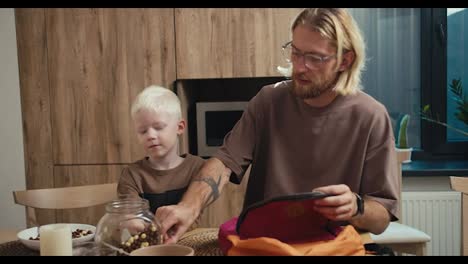 Un-Hombre-Rubio-Con-Gafas-Y-Barba-Ayuda-A-Su-Pequeño-Hijo-Albino-De-Pelo-Blanco-A-Guardar-Cosas-En-Una-Mochila-De-Colores-Brillantes-En-La-Cocina-Después-Del-Desayuno,-Antes-Del-Comienzo-De-La-Jornada-Escolar.