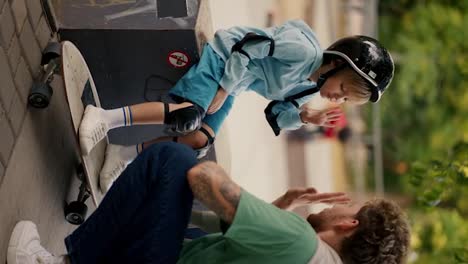 Vertical-video:-Little-blond-boy-in-blue-clothes-in-protective-clothing-for-riding-a-skateboard-in-a-black-helmet-high-fives-his-dad-with-curly-hair-in-a-Green-T-shirt-in-a-skate-park-in-summer