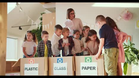 Los-Niños-De-Un-Club-De-Preparación-Escolar-Colocan-Diferentes-Tipos-De-Basura-En-La-Caja-Adecuada-Para-Plástico,-Vidrio-Y-Papel-Junto-Con-Su-Maestra,-Una-Niña-Con-Un-Corte-De-Pelo-Corto-Y-Una-Camisa-Blanca.