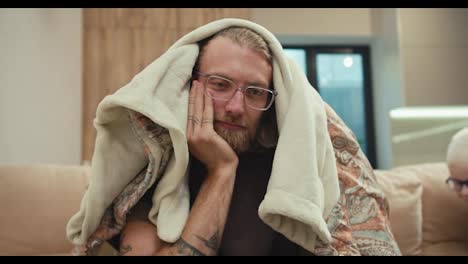 A-tired-blond-man-with-a-beard-and-glasses-covered-himself-with-a-blanket-and-his-little-albino-son-pulls-the-blanket-off-him-and-wants-to-play-with-his-father-in-a-modern-apartment-at-home