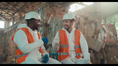 A-man-with-Black-skin-and-a-beard-in-a-white-protective-uniform-sits-and-communicates-with-his-colleague-a-brunette-man-with-a-beard-and-plays-rock-paper-scissors-near-huge-piles-of-garbage-at-a-recycling-plant-and-waste-paper