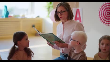 A-blonde-woman-with-a-bob-hairstyle-in-glasses-and-a-white-and-pink-shirt-reads-a-green-book-for-students-in-a-club-for-preparing-children-for-school