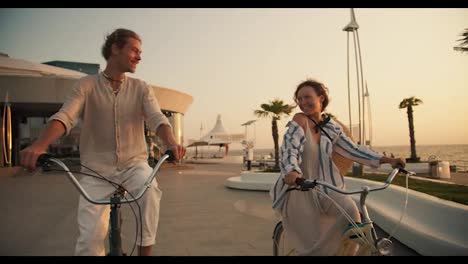 Happy-guy-and-girl-riding-bicycles-on-the-beach-looking-at-each-other-and-holding-hands-near-the-sea-at-sunrise-in-summer.-Happy-couple-ride-bicycles-on-the-beach-with-palm-trees-near-the-sea-in-summer