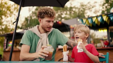 A-father-with-curly-hair-in-a-Green-T-shirt-drinks-a-cocktail-and-his-little-blond-boy-with-blue-eyes-in-a-red-T-shirt-eats-hot-dogs-holding-them-in-two-hands-while-sitting-at-a-table-in-a-street-cafe-in-the-park