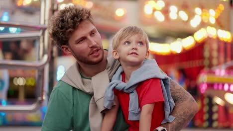 A-little-blond-boy-in-a-red-T-shirt-sits-on-his-dad-s-leg-and-together-they-look-at-the-bright-and-colorful-attractions-in-the-amusement-park-and-communicate